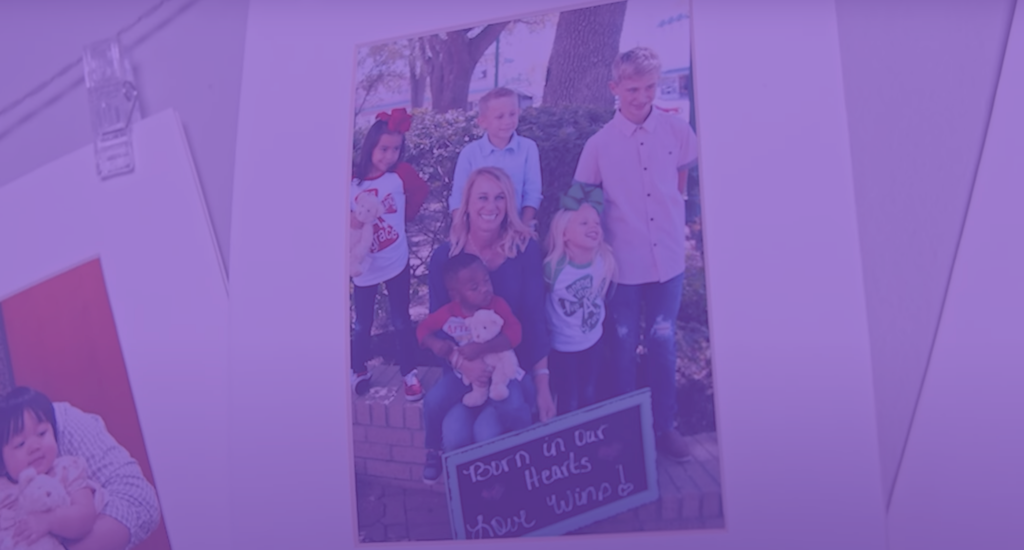 A photo of a lady sitting with 5 kids, holding a sign that say, born in our hearts. Love wins!